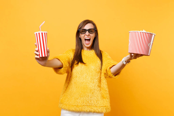 Crazy young woman in 3d imax glasses screaming, watching movie film, holding bucket of popcorn, plastic cup of cola or soda isolated on yellow background. People sincere emotions in cinema, lifestyle - Valokuva, kuva