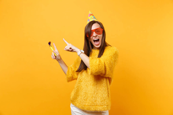 Opgewonden gelukkige vrouw in oranje grappige bril, verjaardag hoed met spelen pijp wijzen wijsvinger opzij op kopie ruimte, vieren geïsoleerd op gele achtergrond. Mensen oprechte emoties, lifestyle - Foto, afbeelding