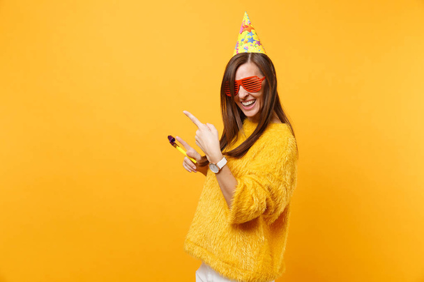 Cheerful happy woman in orange funny glasses, birthday hat with playing pipe pointing index fingers aside on copy space, celebrating isolated on yellow background. People sincere emotions, lifestyle - Photo, Image