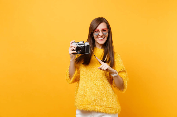 Portrait of smiling young woman in heart glasses pointing index finger on retro vintage photo camera isolated on bright yellow background. People sincere emotions, lifestyle concept. Advertising area - Photo, Image