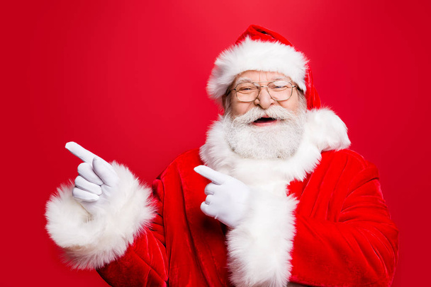 Funky rejoice stylish Santa in costume gloves headwear and spectacles with white beard indicate aside forefinger both hands show presentation product promotion isolated on noel red background - Fotó, kép