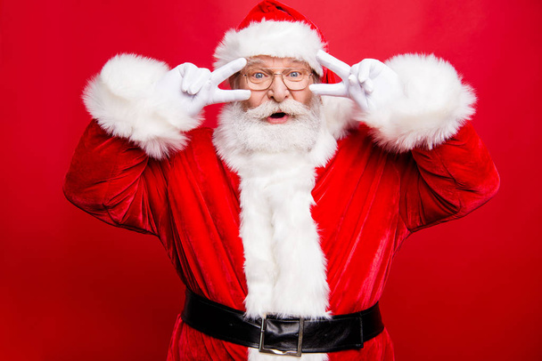Portrait of positive aged trendy Santa in costume gloves and headwear look at camera open mouth staring eyes show v-sign by fingers near face isolated on shine red background - Zdjęcie, obraz