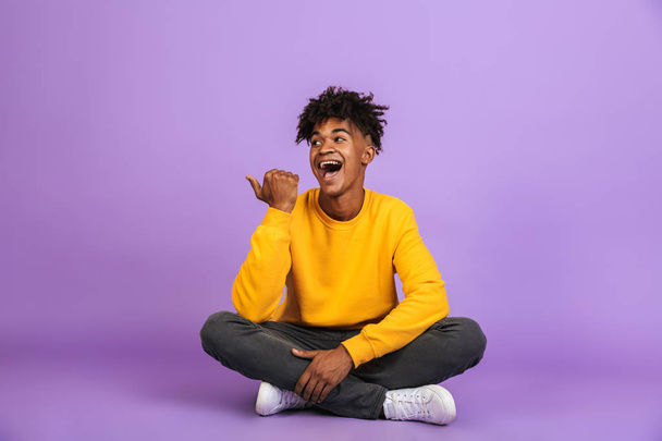 Portrait of happy african american guy smiling and pointing finger aside at copyspace while sitting on floor with legs crossed isolated over violet background - Фото, зображення