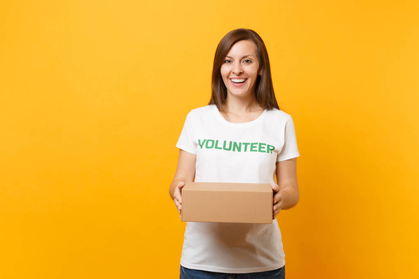 Porträt einer lächelnden Frau in weißem T-Shirt mit Schriftzug Grüner Titel Freiwilliger mit leerem Pappkarton auf gelbem Hintergrund. Freiwillige, kostenlose Hilfe, Wohltätigkeitskonzept - Foto, Bild