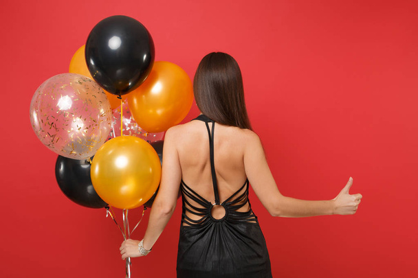 Back rear view of young girl in little black dress celebrating showing thumb up hold air balloons isolated on red background. St. Valentine's Day Happy New Year, birthday mockup holiday party concept - Photo, Image