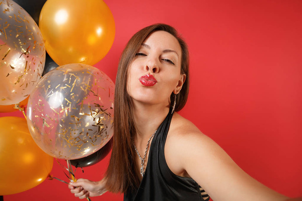 Primer plano selfie disparo de mujer en vestido negro celebrando soplado labios beso mantenga globos de aire aislados sobre fondo rojo. Día Internacional de la Mujer Feliz Año Nuevo concepto de fiesta simulada de cumpleaños
 - Foto, Imagen