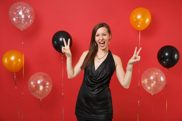 Jovencita alegre en pequeño vestido negro celebrando, mostrando signo de victoria en globos de aire de fondo rojo brillante. San Valentín, Día de la Mujer, Feliz Año Nuevo, concepto de fiesta de cumpleaños
 - Foto, imagen