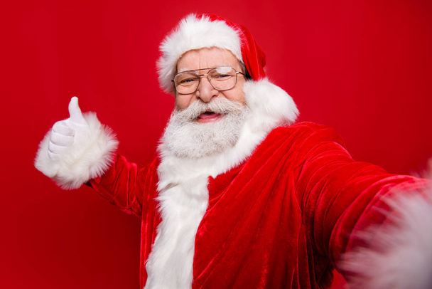 Aged mature stylish grandfather Santa Claus with white beard in costume headwear gloves stylish spectacles show like sign thumb up take picture on camera of smartphone isolated on red background - Fotó, kép