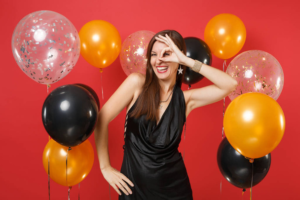 Γέλιο νεαρή γυναίκα στο μικρό μαύρο φόρεμα γιορτάζει, δείχνει εντάξει σημάδι κοντά τα μάτια σε λαμπερό κόκκινο φόντο και αερόστατα. Του Αγίου Βαλεντίνου ημέρα, ευτυχισμένο το νέο έτος, γενέθλια mockup holiday κόμμα έννοια - Φωτογραφία, εικόνα