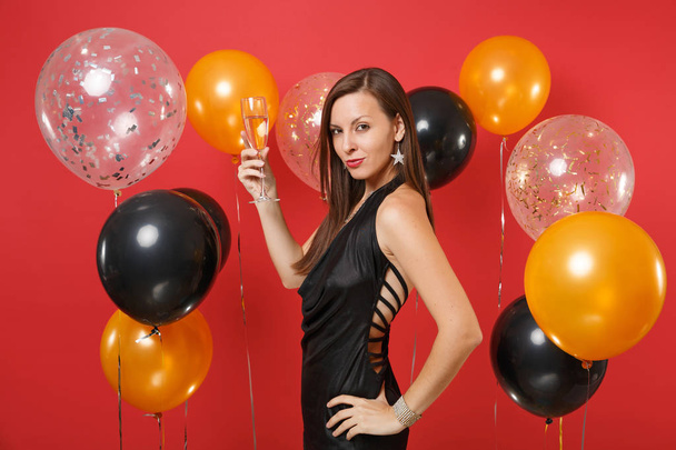 Side view of passionate girl in black dress celebrating, holding glass of champagne on bright red background air balloons. St. Valentine's Day, Happy New Year, birthday mockup holiday party concept - Photo, Image