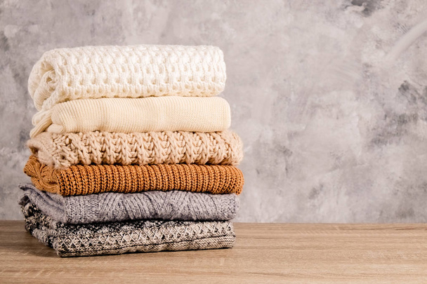 Bunch of knitted warm pastel color sweaters with different knitting patterns folded in stack on brown wooden table, grunged concrete wall background. Fall winter season knitwear. Close up, copy space - Foto, imagen