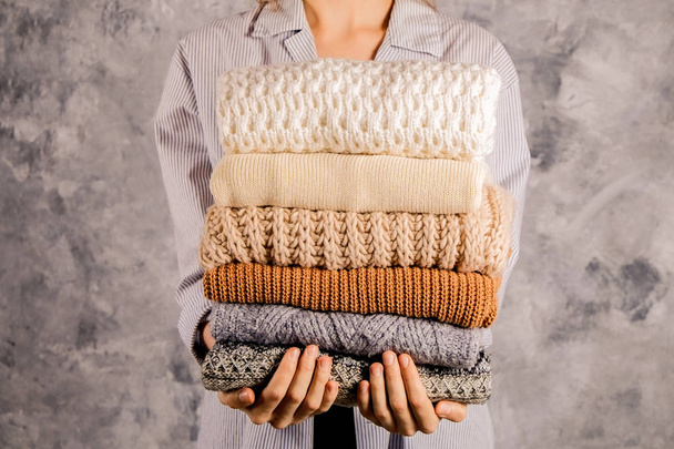 Slim young woman wearing blue oversized unbuttoned cotton shirt and holding big stack of folded knitted warm pastel color sweaters, easy chic style, different knitting patterns. Background, copy space - Photo, Image