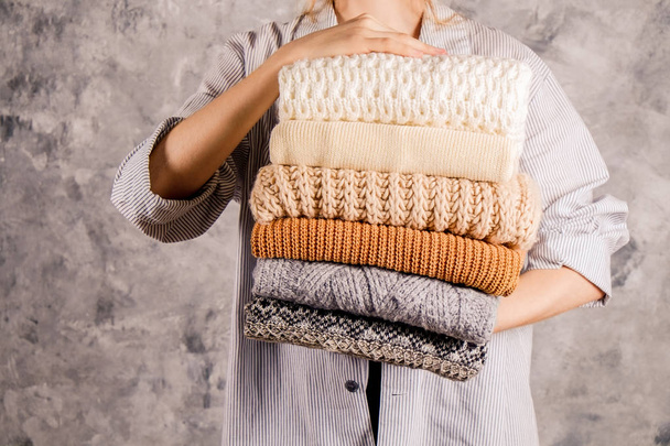Slim young woman wearing blue oversized unbuttoned cotton shirt and holding big stack of folded knitted warm pastel color sweaters, easy chic style, different knitting patterns. Background, copy space - 写真・画像