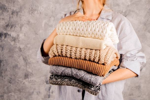 Slim young woman wearing blue oversized unbuttoned cotton shirt and holding big stack of folded knitted warm pastel color sweaters, easy chic style, different knitting patterns. Background, copy space - Photo, Image