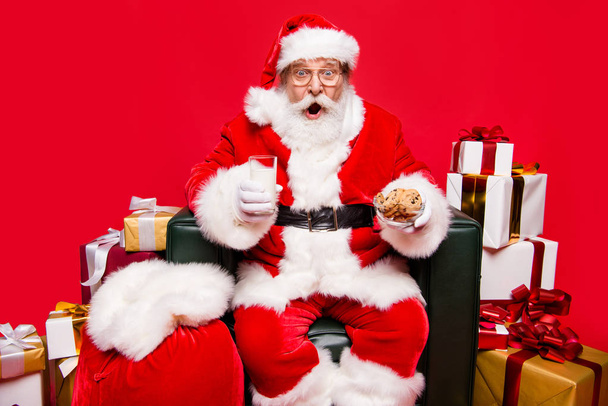 Fun joy winter wish December noel mature aged Nicholas in headwear white beard isolated on red shine background open mouth staring eyes unbelievable tasty yummy homemade chocolate pastry sit on chair - Foto, immagini
