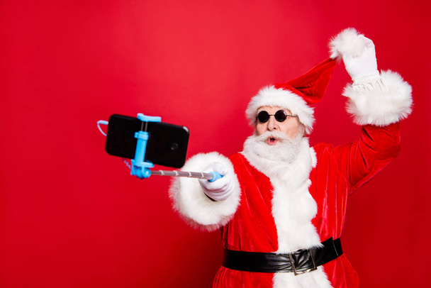 Winter December noel christmastime eve wish trendy stylish aged mature senior Santa headwear tradition costume white beard take selfie picture on front camera smartphone isolated on red background - 写真・画像