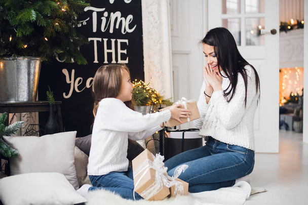 Счастливого Рождества и счастливых праздников. Веселая мама и ее милая дочка открывают рождественский подарок. Родители и маленькие дети веселятся возле рождественской елки в помещении - Фото, изображение
