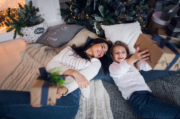 Merry Christmas and Happy Holidays. Cheerful mom and her cute daughter girl exchanging gifts. Parent and little child having fun near Christmas tree indoors. Loving family with presents in room. - Photo, Image