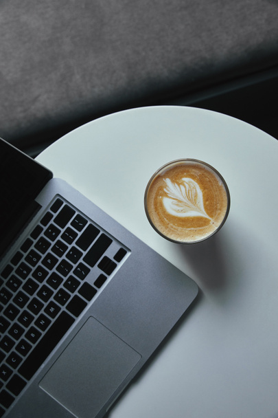 κάτοψη του φλυτζάνι του cappuccino και φορητό υπολογιστή στο τραπέζι στην καφετέρια  - Φωτογραφία, εικόνα