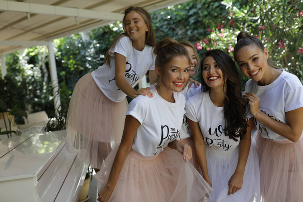 Mujeres jóvenes activas en las faldas de color rosa claro y camisetas blancas con el título de pie cerca del piano, terraza, evento, fiesta, maquillaje, peinado, moda, belleza, risa, sonrisa, loco
 - Foto, Imagen