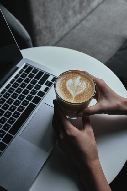 partial view of person holding cup with cappuccino and using laptop - Φωτογραφία, εικόνα