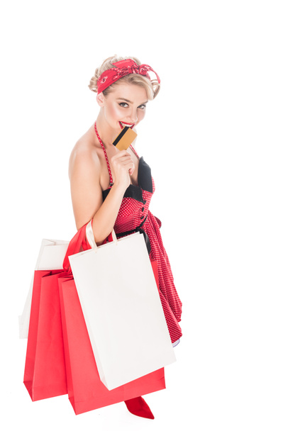 high angle view of woman in pin up clothing with shopping bags biting credit card isolated on white - Fotó, kép