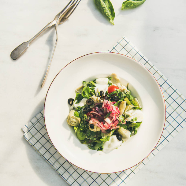 Fresh green summer salad with artichokes, olives, soft cheese and red onion in white plate over light marble table background - Foto, Bild