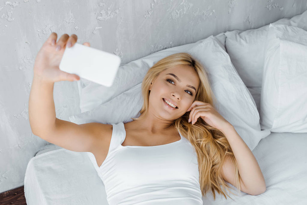 vista de ángulo alto de la joven sonriente acostada en la cama y tomando selfie por teléfono inteligente
 - Foto, Imagen