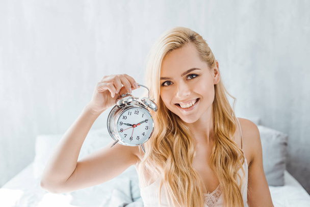 beautiful young blonde woman holding alarm clock and smiling at camera in bedroom - Zdjęcie, obraz