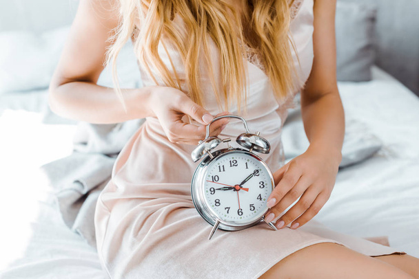 cropped shot of young blonde woman sitting on bed and holding alarm clock - Foto, Imagen
