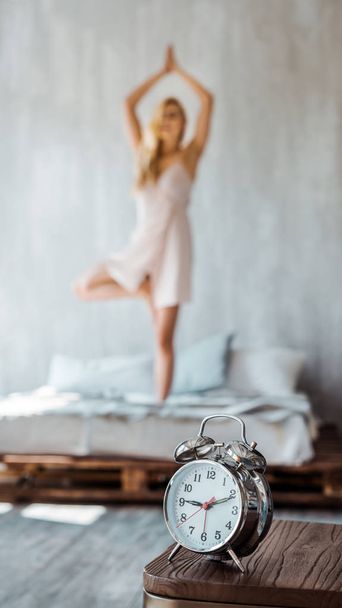 vergrote weergave van de wekker op houten tafel en jonge vrouw uitvoeren van yoga op bed achter  - Foto, afbeelding