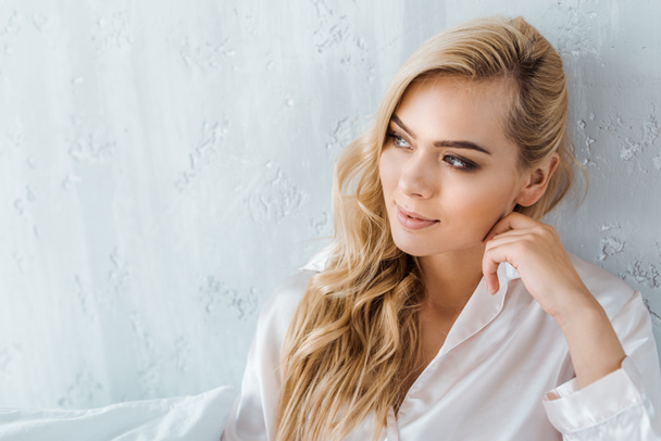 beautiful smiling young blonde woman in pajamas sitting on bed and looking away - 写真・画像
