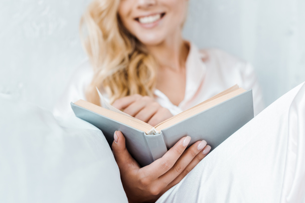close-up view of happy young woman in pajamas sitting and reading book - Фото, изображение