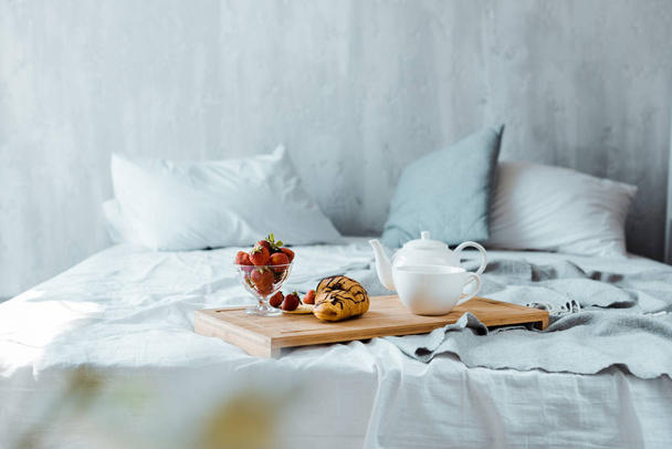 Croissant, aardbeien en thee op houten dienblad op bed - Foto, afbeelding