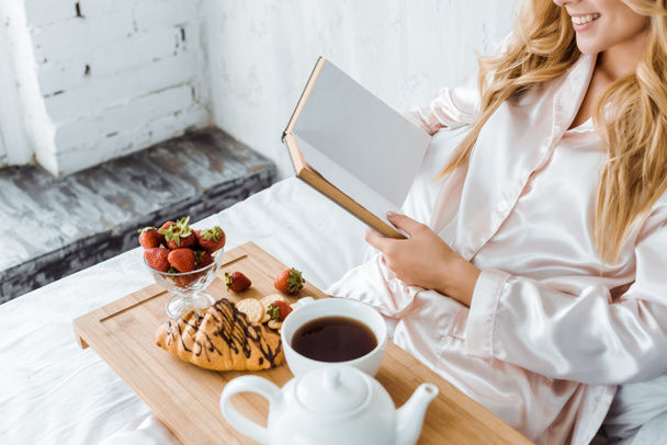 immagine ritagliata di donna attraente in pigiama libro di lettura, colazione su vassoio di legno a letto
 - Foto, immagini