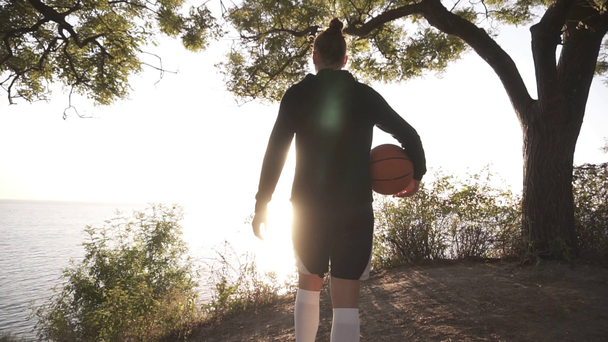 Баскетболистка приходит с мячом в руке, подходя к склону с деревьями вокруг. Смотрит на солнце, сияющее над морем. Вид сзади
 - Кадры, видео