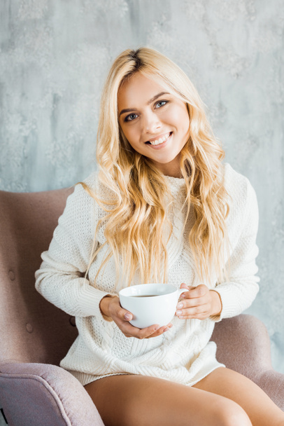 smiling attractive woman in sweater sitting on armchair and holding cup of coffee in bedroom in morning - Фото, изображение