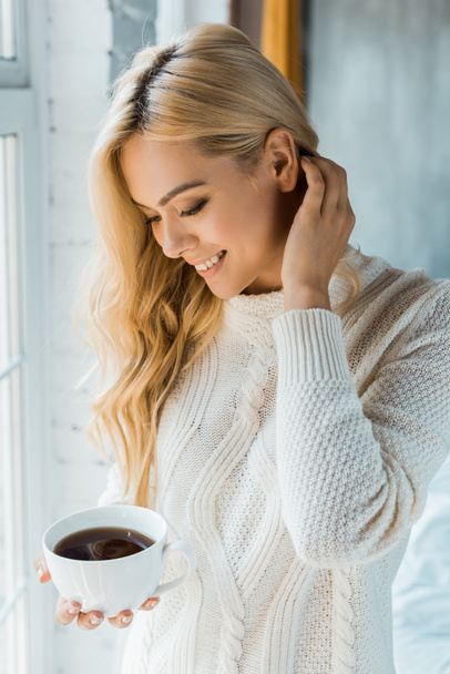 セーターの寝室で朝の一杯のコーヒーを保持している笑顔の魅力的な女性 - 写真・画像