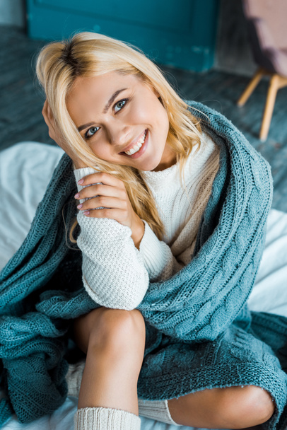 smiling attractive woman in sweater sitting wrapped in blanket and looking at camera in bedroom in morning - Valokuva, kuva