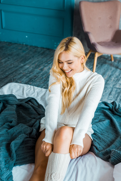 lächelnde attraktive Frau im Pullover mit gestrickten Gamaschen morgens im Schlafzimmer - Foto, Bild