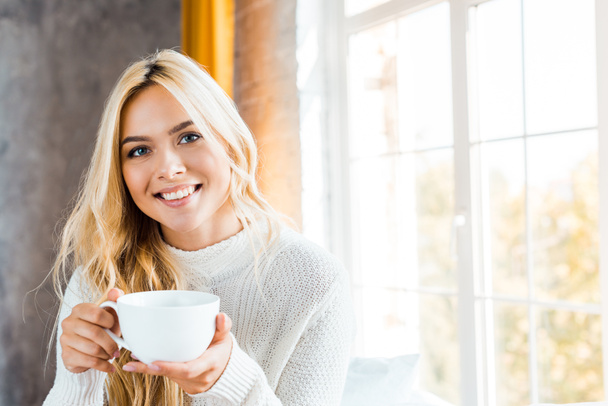 cheerful beautiful woman in sweater holding cup of coffee and looking at camera in bedroom in morning - Fotografie, Obrázek