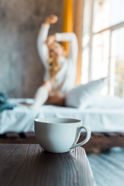 selective focus of attractive woman in sweater stretching in bedroom in morning, cup of coffee on tabletop - Foto, Bild