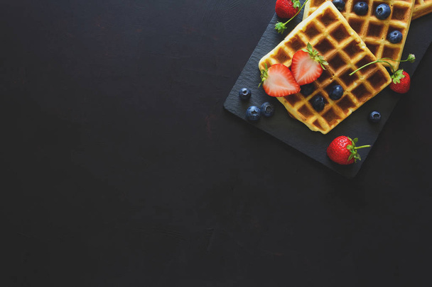 Belgian waffles with strawberries and blueberries on slate plate on dark wooden background. Top view. Copy space - Φωτογραφία, εικόνα