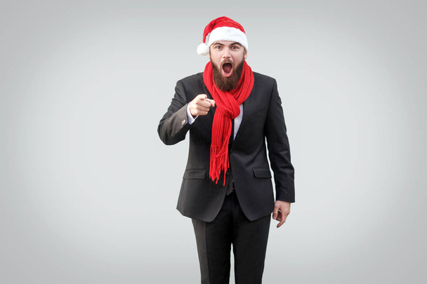 Surprised handsome bearded businessman in black classic suit and christmas cup with red scarf looking and pointing at camera on gray background - Foto, immagini