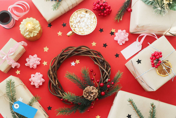 Kerst krans en giften onder decoratie naast een kopje chocolademelk met marshmallows op een rode achtergrond. Bovenaanzicht, plat lag - Foto, afbeelding
