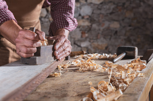 Carpinteiro alisando feixe de madeira longa em cima da mesa com plano de mão e outras tábuas na frente da parede de pedra
 - Foto, Imagem