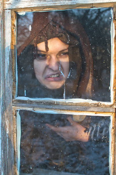 молодая ведьма корчится чудовищно, полна яростного гнева отвратительной гримасы смотрит в окно своей хижины явно показывая отвращение и отвращение к миру
 - Фото, изображение