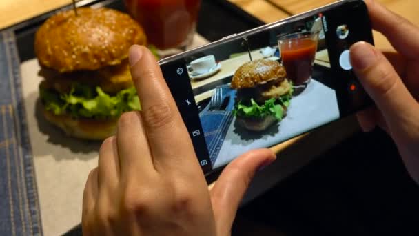 Menina faz uma foto de hambúrguer e suco de tomate em um smartphone em um café de perto
 - Filmagem, Vídeo