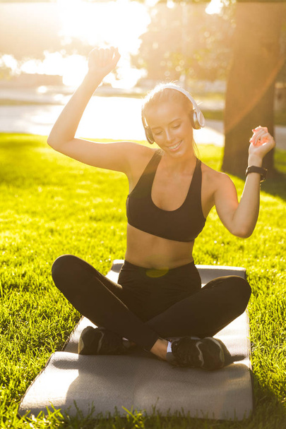 Photo of beautiful young blonde sports woman in park outdoors listening music with headphones. - 写真・画像