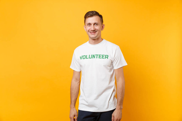 Muotokuva onnellinen hymyilevä tyytyväinen nuori mies valkoinen t-paita kirjallisella kirjoitus vihreä otsikko vapaaehtoinen eristetty keltaisella pohjalla. Vapaaehtoinen maksuton apu apua, hyväntekeväisyys armo työtä käsite
 - Valokuva, kuva